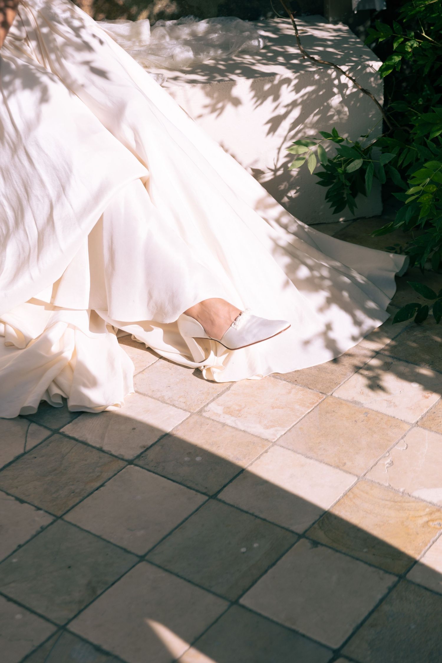White wedding dress hem and bridal heels on sunlit stone tiles with green foliage shadows.