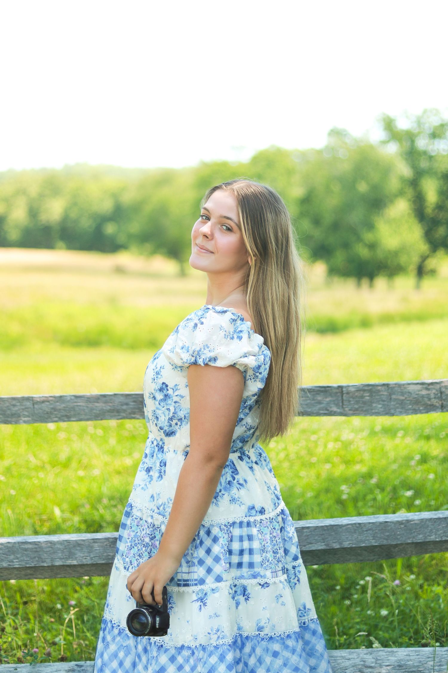 Young woman in blue floral dress holding camera, standing by wooden fence in sunny green field.