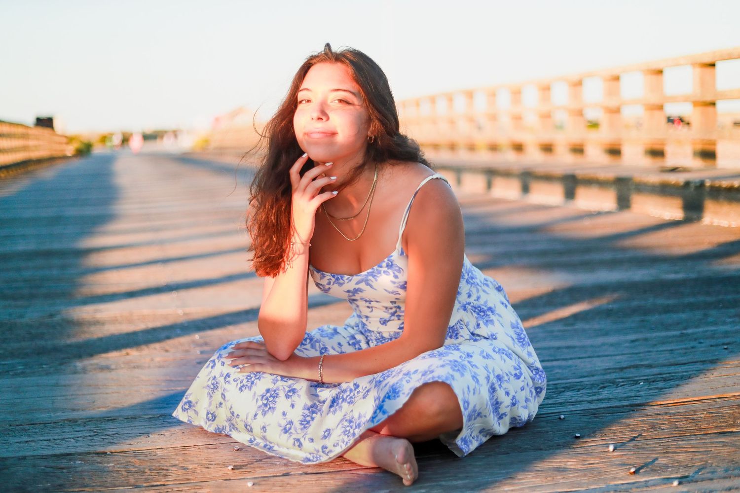 A woman in a floral dress poses at sunset on a wooden boardwalk.