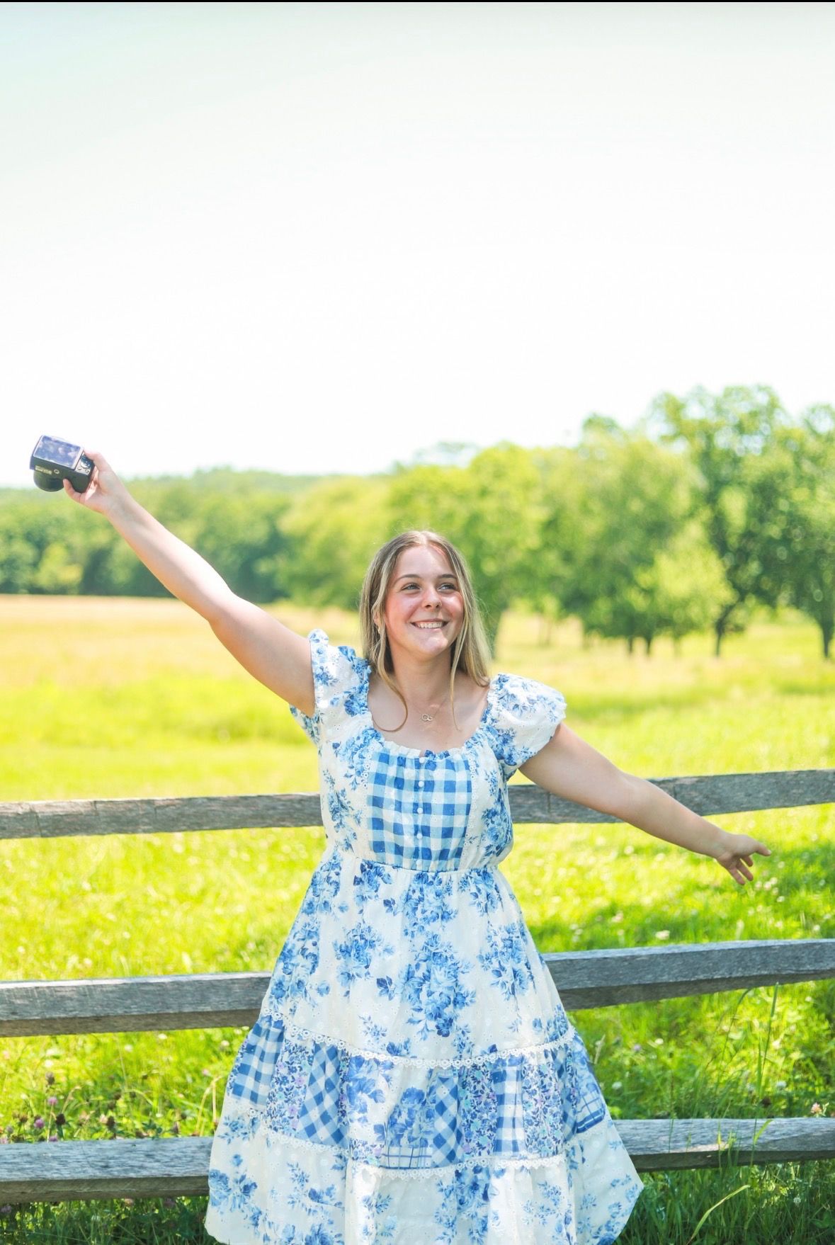 Young woman in blue floral dress holding camera, arms outstretched, smiling outdoors by wooden fence in green field.