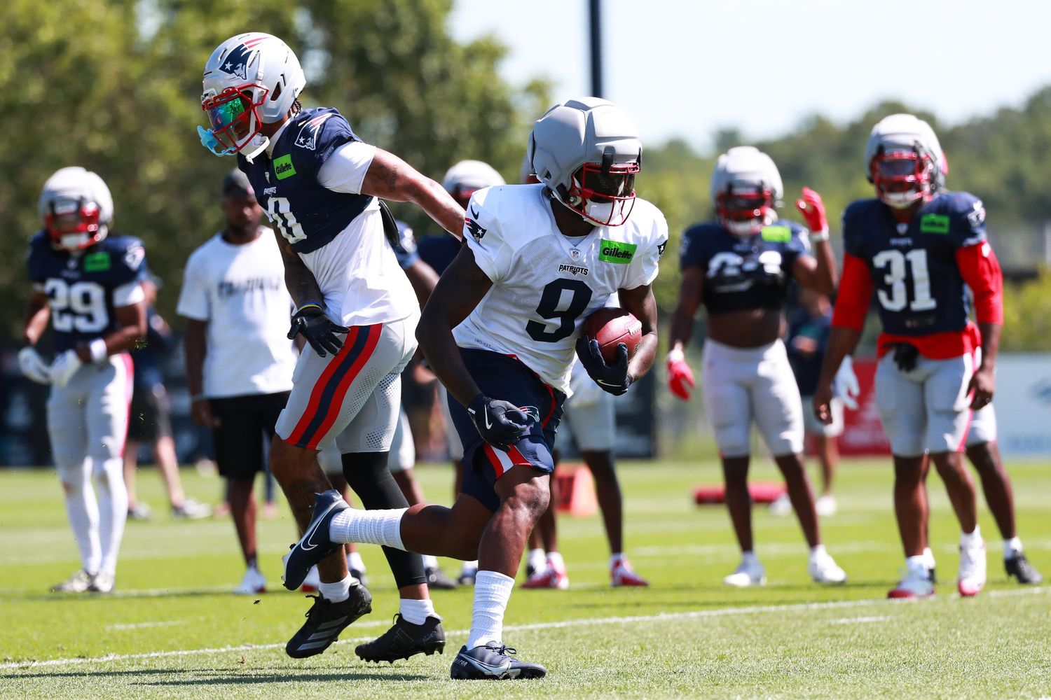 NFL players in Patriots uniforms practice drills on a sunny green field during training camp.