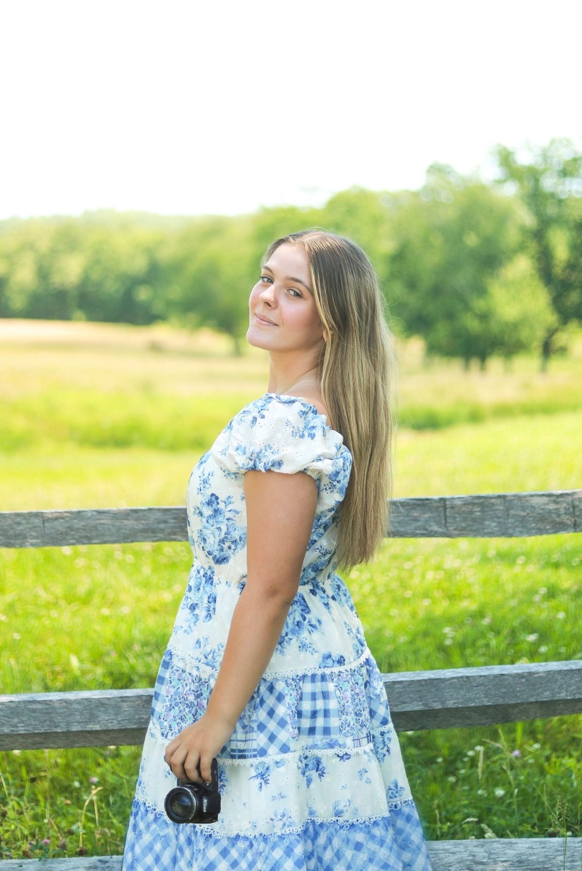 Young woman in blue floral dress holding camera, standing by wooden fence in sunny green field.