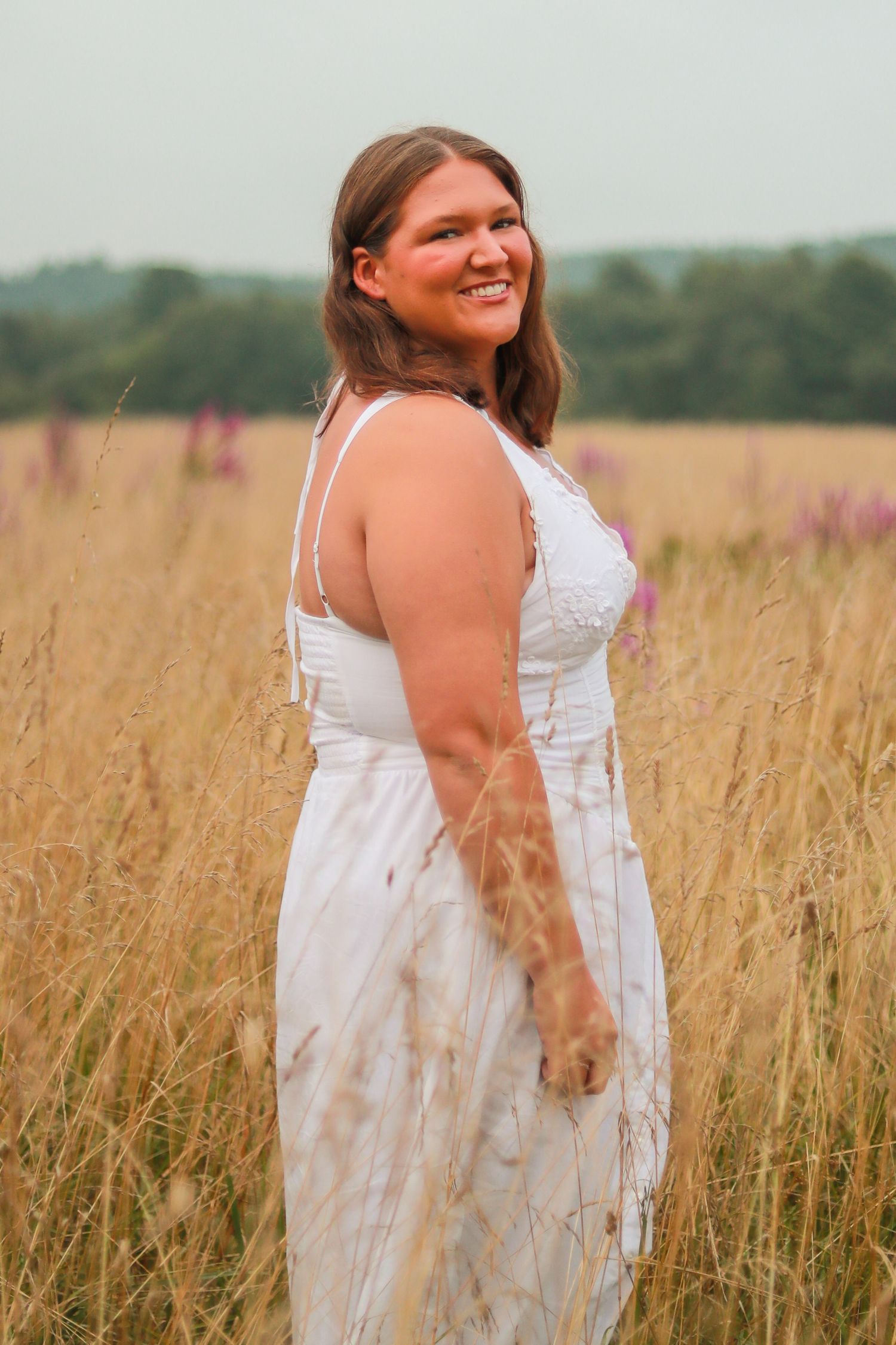 A woman wearing a white dress stands in a golden wheat field during sunset.