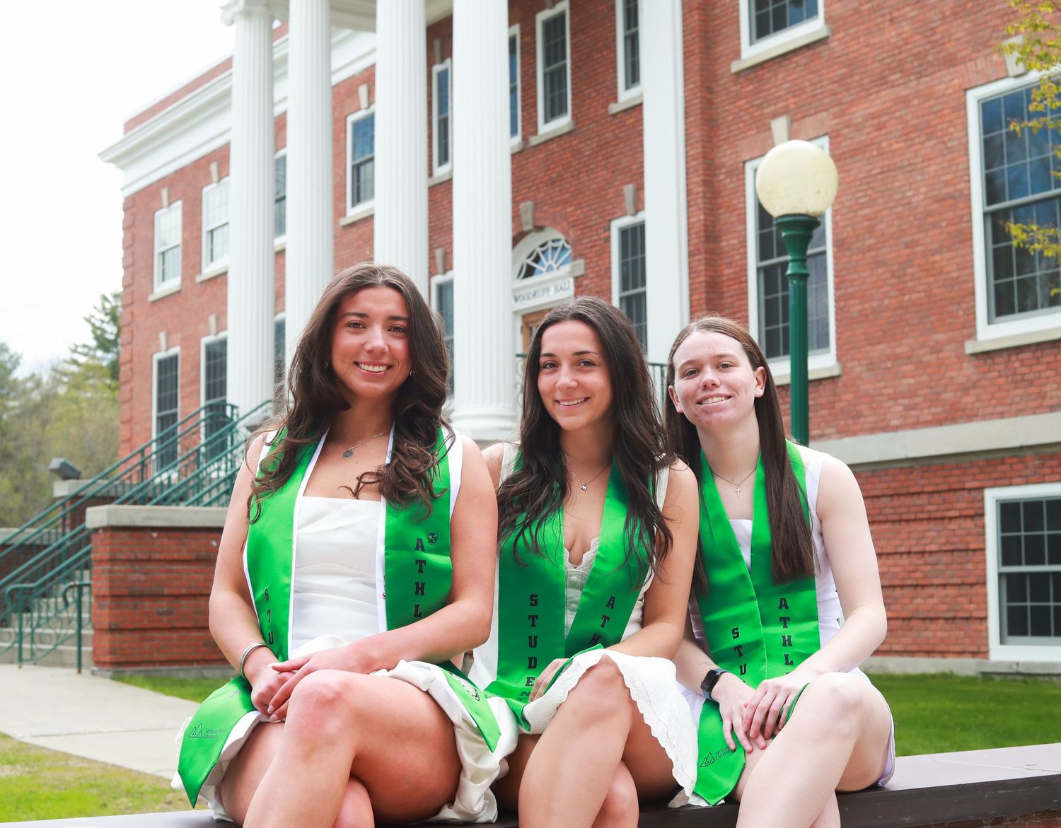Three young women in green graduation sashes smile together outdoors in front of a brick building on a sunny day.