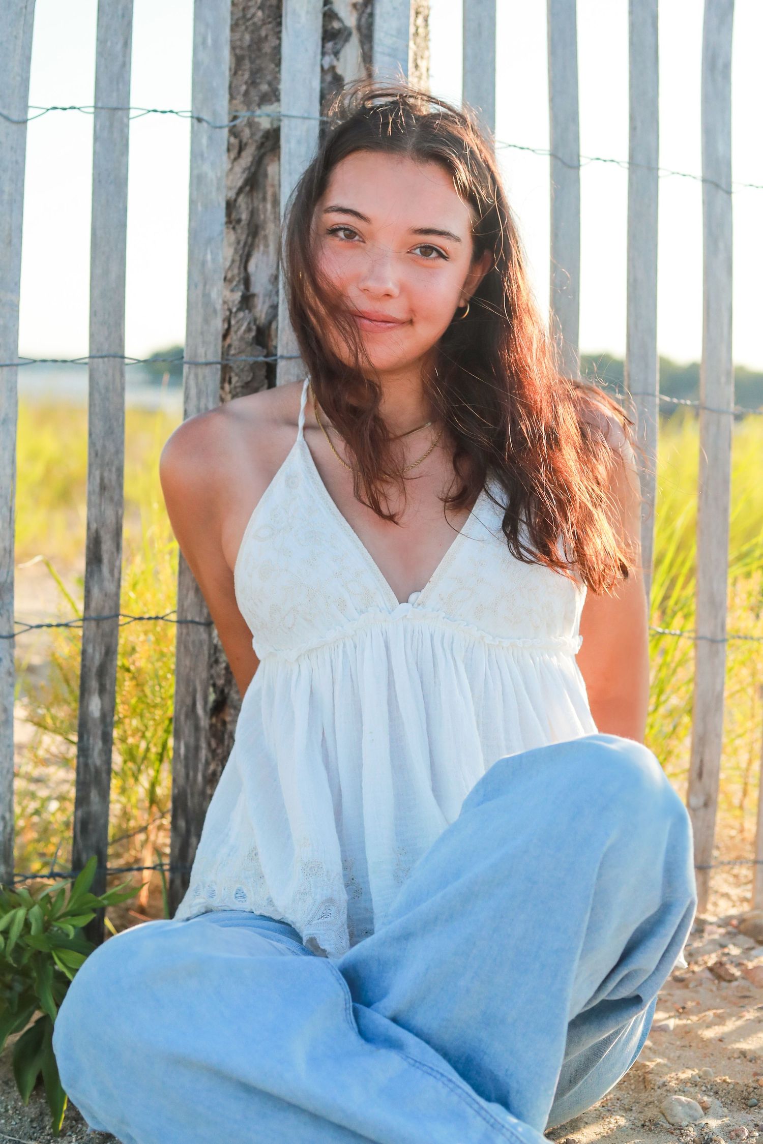 A person in white top and blue jeans sits casually against white fence posts.