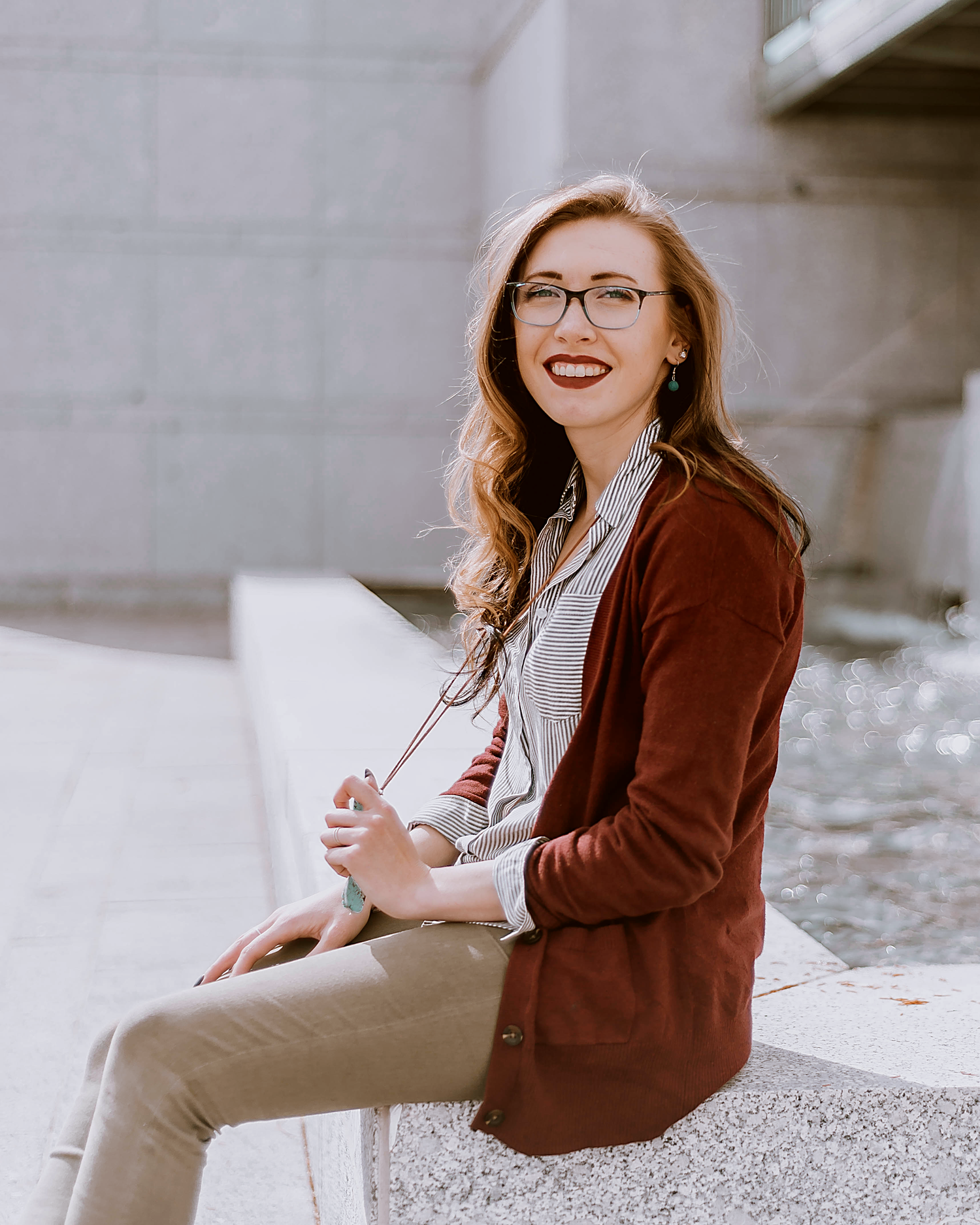 Professional wearing glasses and brown cardigan sits outdoors in natural lighting for a casual portrait.