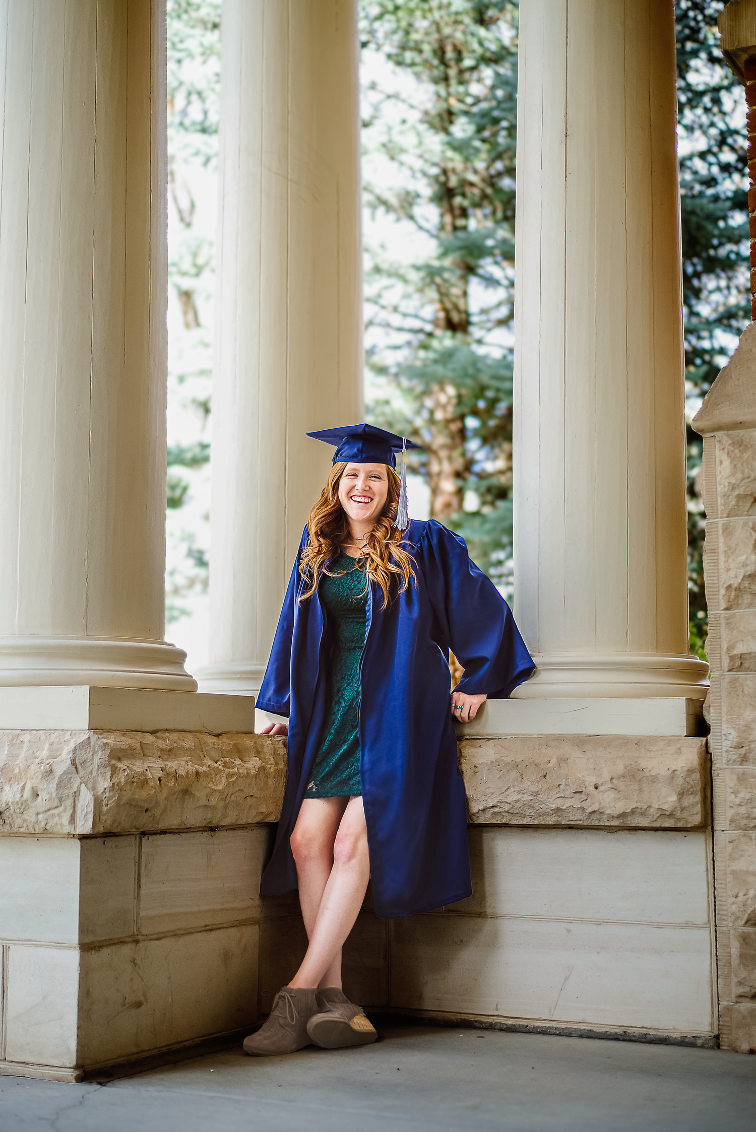 Graduate in blue cap and gown poses between elegant white columns at graduation ceremony.