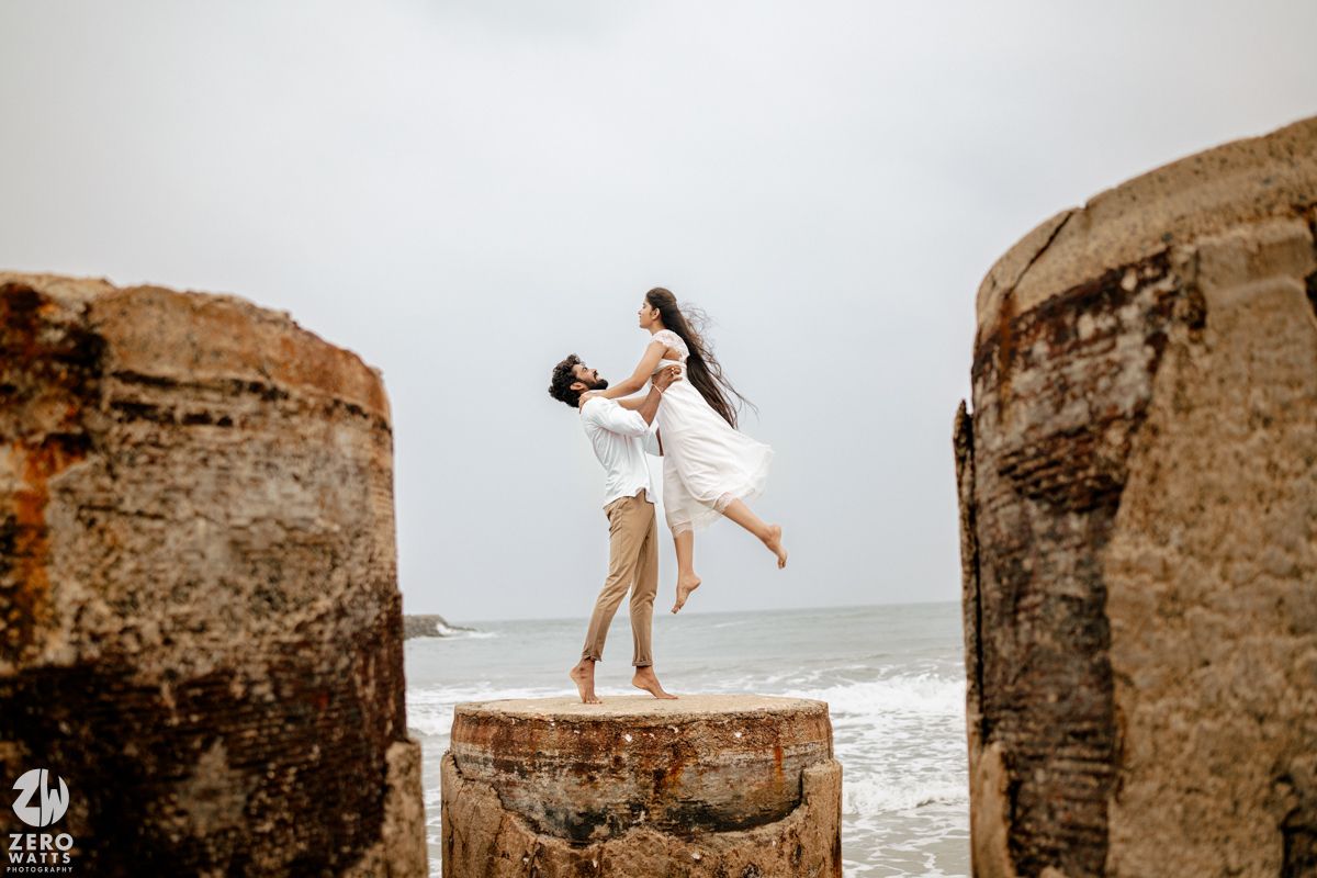 Romantic couple sharing a lift pose between ancient stone pillars with ocean waves in the background.