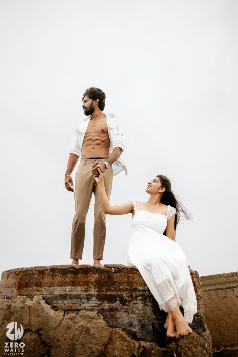 Couple on rocky ocean structure, man in open white blazer standing holding hand of woman in white dress.