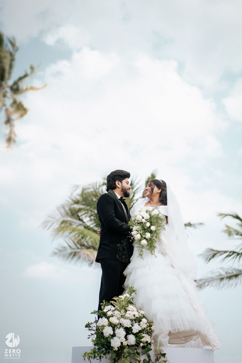 Bride and groom in elegant wedding attire stand on elevated platform with palm trees and blue sky backdrop.