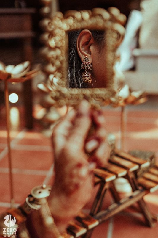 Ornate gold-framed mirror reflecting South Indian bride wearing traditional jhumka earrings with mehndi hands.