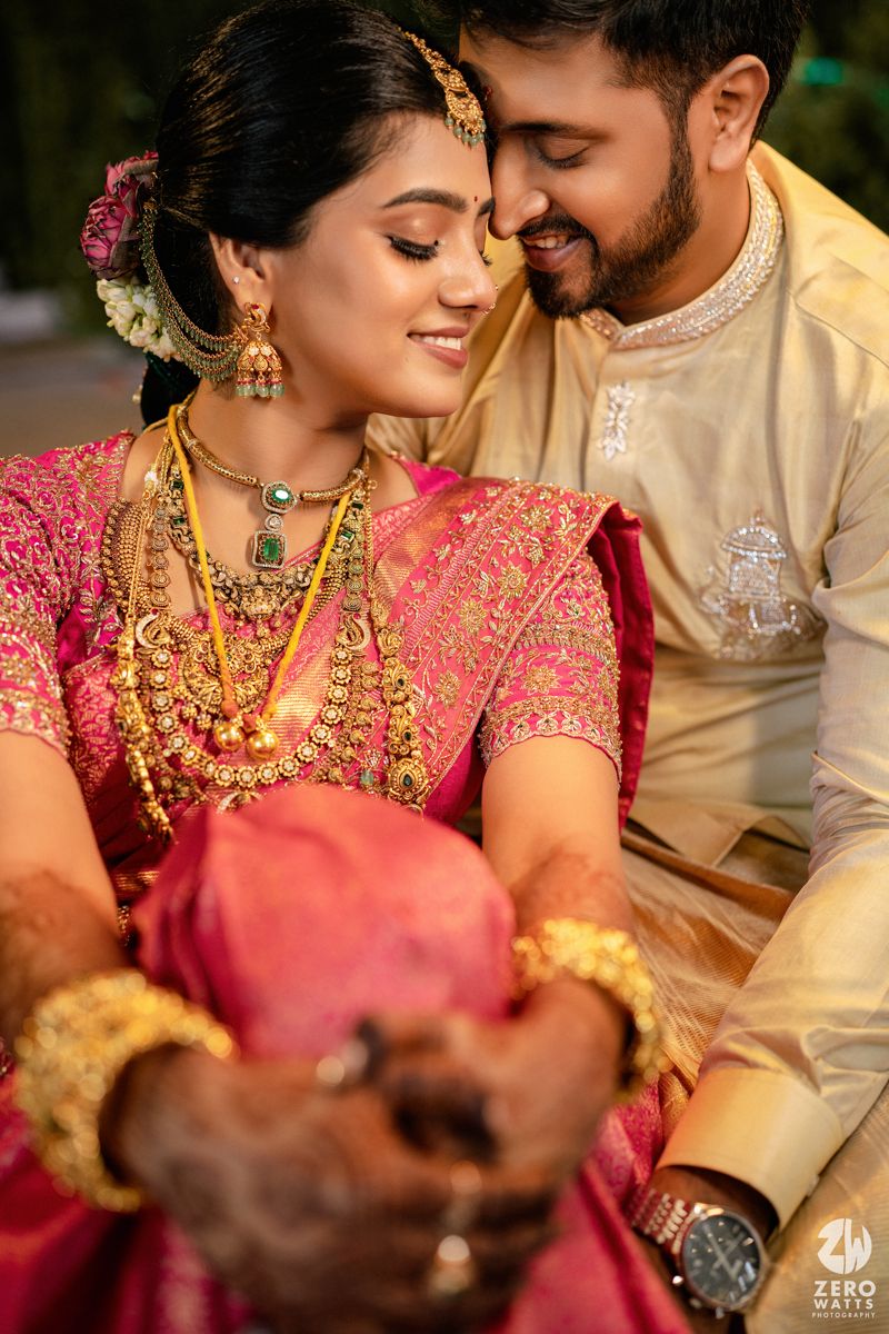 South Indian bride in pink silk saree adorned with gold jewelry shares intimate moment with groom in cream sherwani.