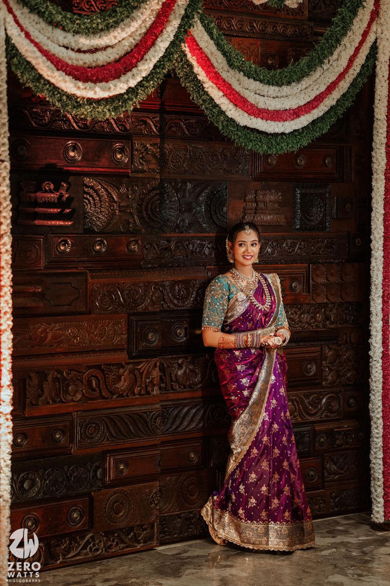 Bride in purple silk saree posing before ornate carved wooden door with floral garlands.