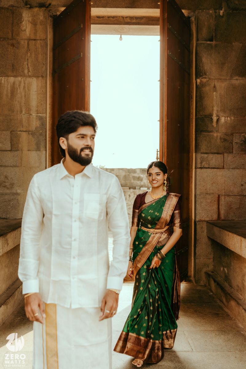 Groom in white kurta and bride in green Kanjivaram saree pose in ancient stone temple doorway.