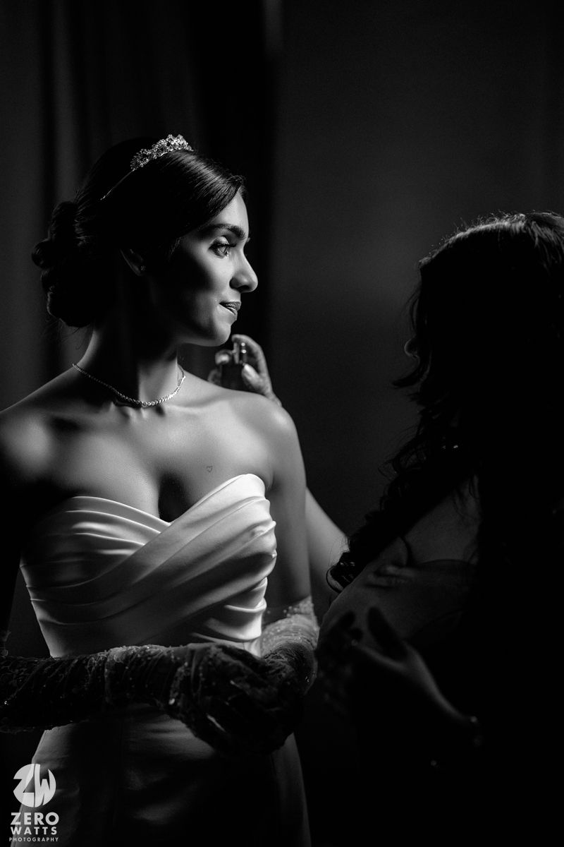 Black and white photo of bride in strapless gown being assisted by attendant.