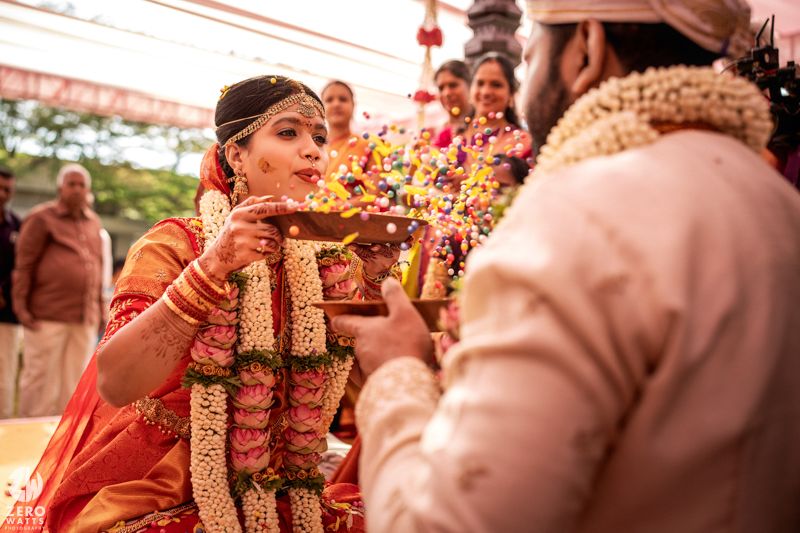 Bride in red saree and groom exchange flowers during traditional South Indian wedding ceremony ritual.