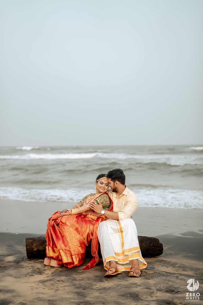 South Indian wedding couple sitting on driftwood log on sandy beach in traditional attire.