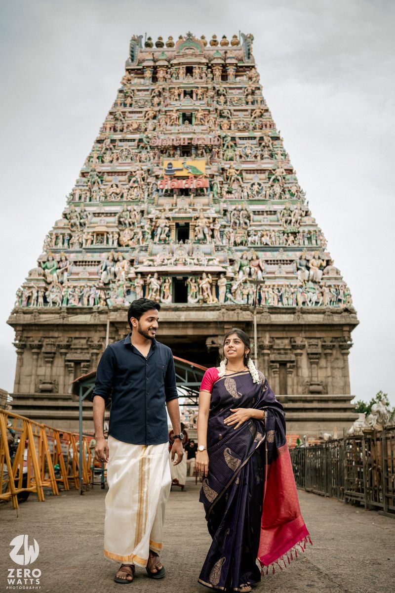 Couple in traditional Indian attire posing against backdrop of ornate temple gopuram on cloudy day.