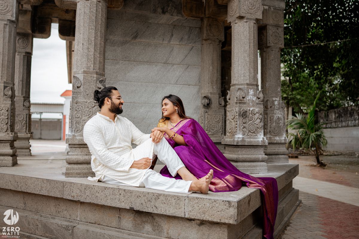 A couple in white and purple attire reclines on a stone ledge of an ornate traditional architectural structure.