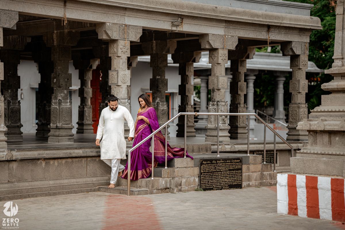 Elegant couple in traditional Indian wedding outfits walking through a temple corridor with stone pillars.