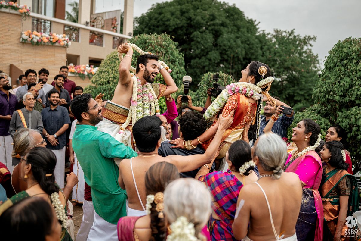 Guests at an outdoor Indian wedding ceremony raise their hands in celebration under lush green trees.
