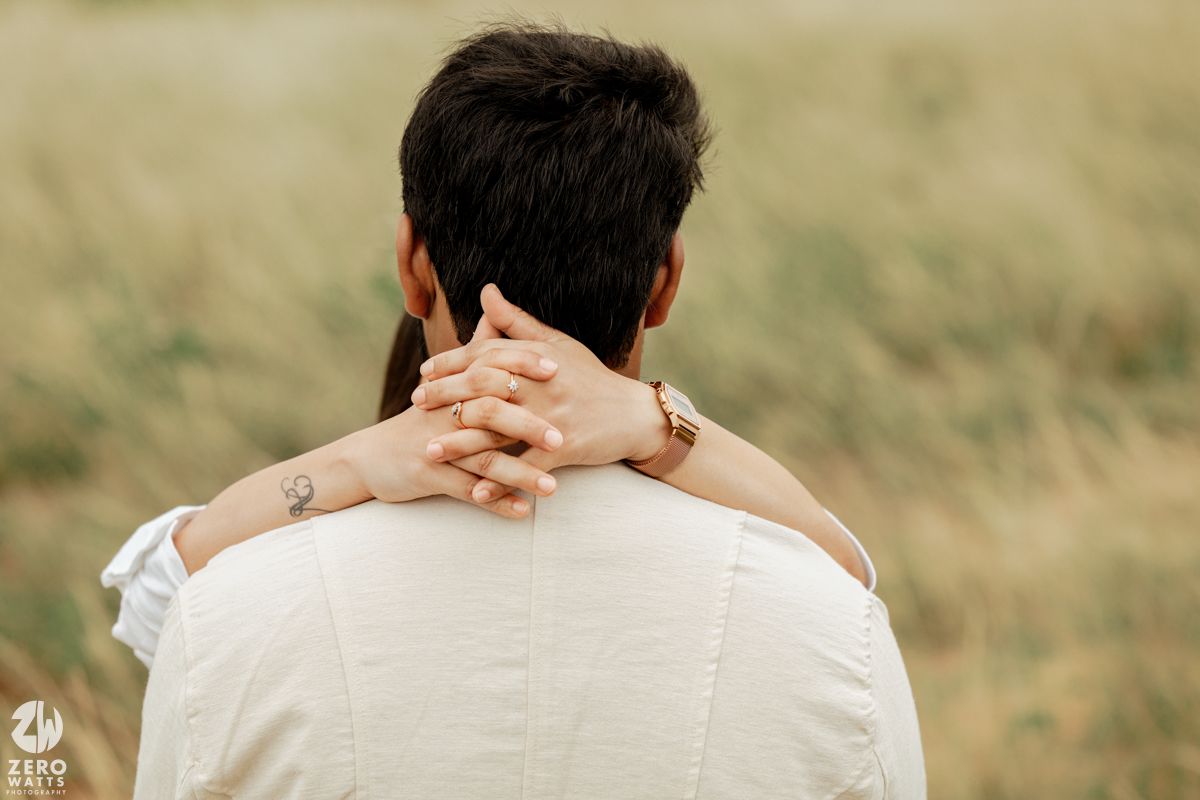 Two people hold hands while sitting together in a grassy outdoor setting during golden hour.