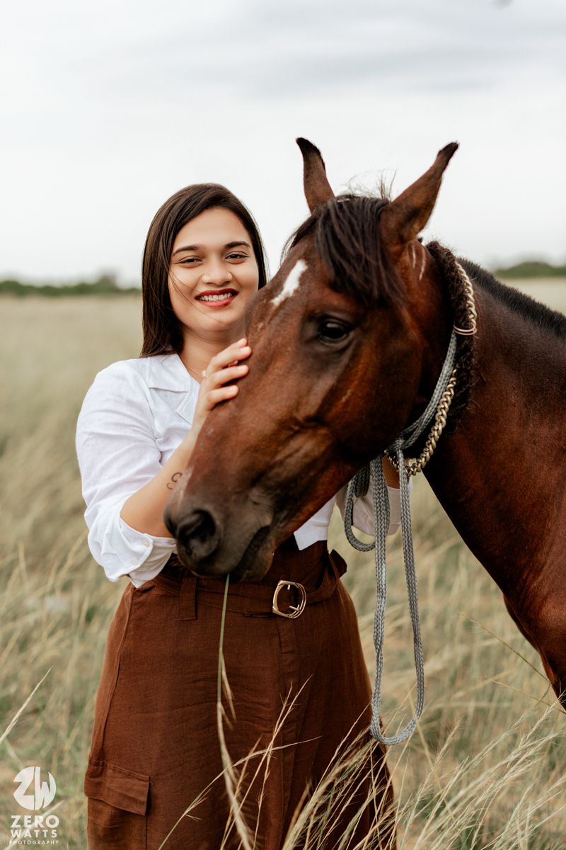 A brown horse with a bridle poses with a person in white against a rustic outdoor backdrop.