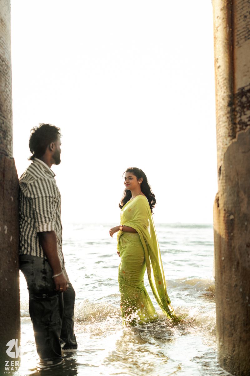 Two people stand between weathered concrete pillars in shallow ocean water during a beach photoshoot.