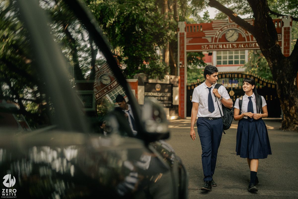 Two students in school uniforms walk together on a tree-lined street near a brick building.