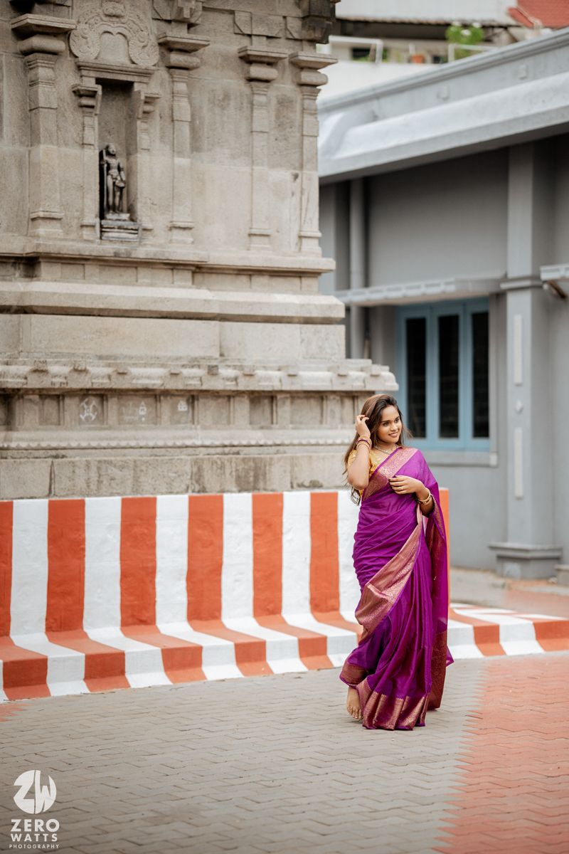 Woman in vibrant purple saree poses elegantly against temple architecture with striped barrier.