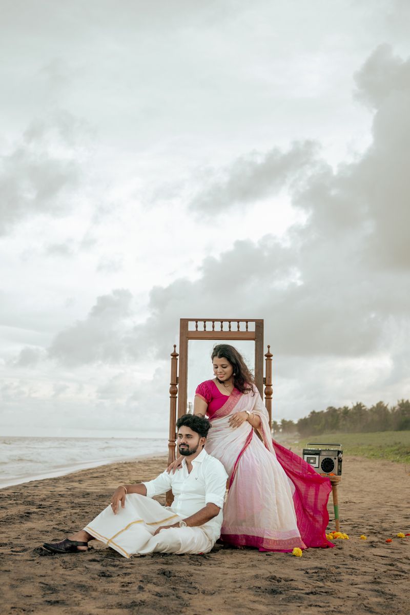 Romantic beach photoshoot with couple lounging on antique wooden chair against cloudy coastal backdrop.