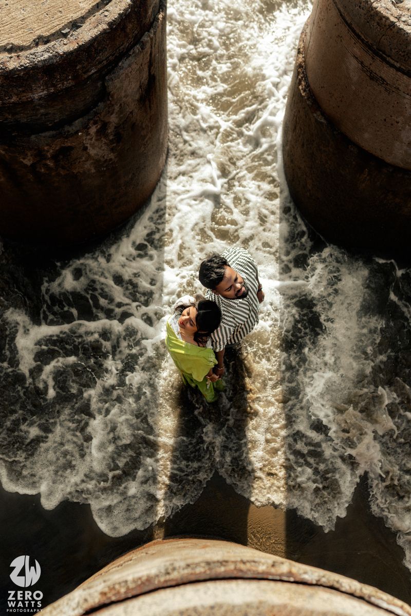Ocean waves surge around concrete pillars with people visible from an aerial perspective.