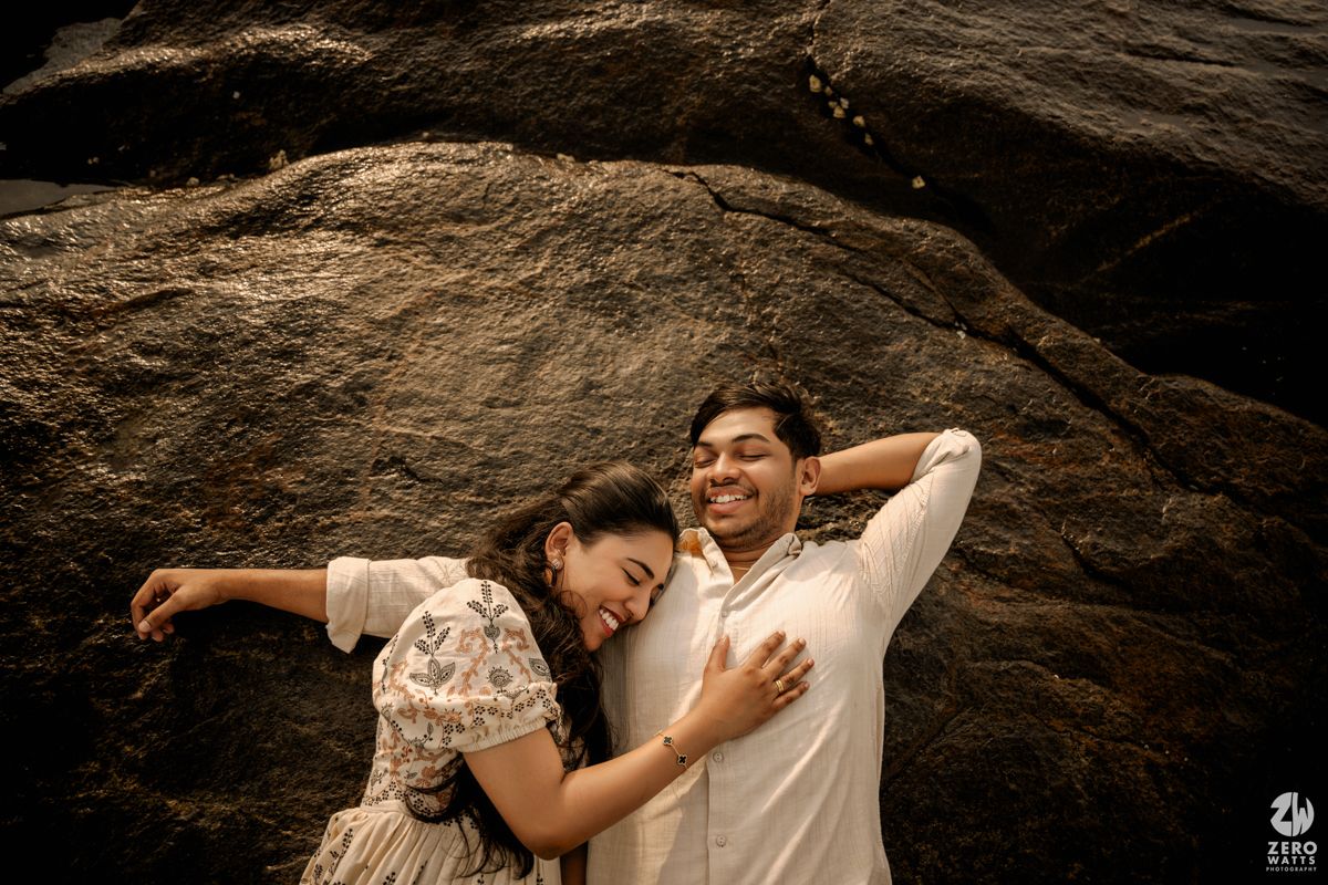 Two people embracing while lying on dark rocky terrain near the ocean.