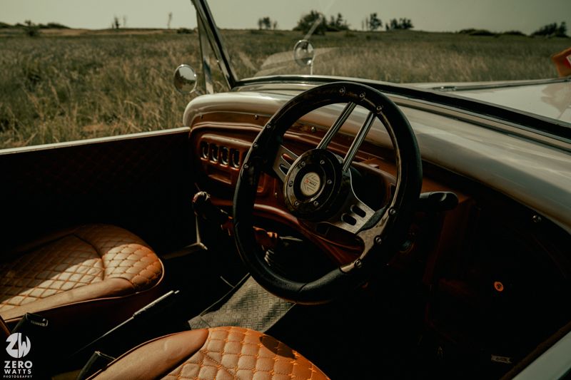 Interior view of a classic car with brown leather quilted seats and wooden steering wheel against rural landscape.