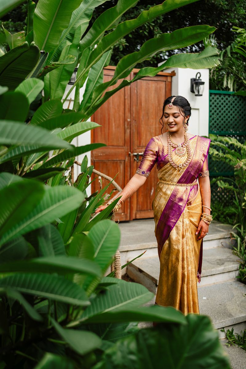 Traditional South Asian attire is showcased in an outdoor garden setting with lush green foliage and wooden fence.
