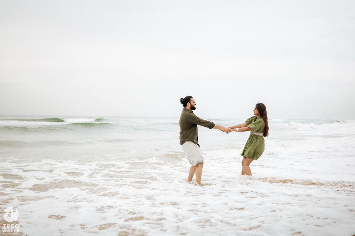 Two people hold hands while standing in the shallow surf at the beach.