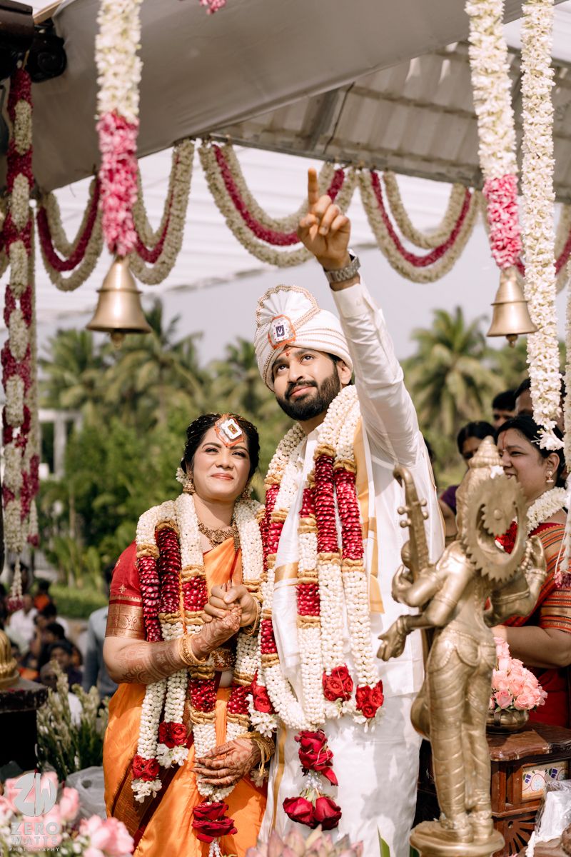 South Indian couple in traditional wedding attire at decorated mandap with floral garlands and bells.
