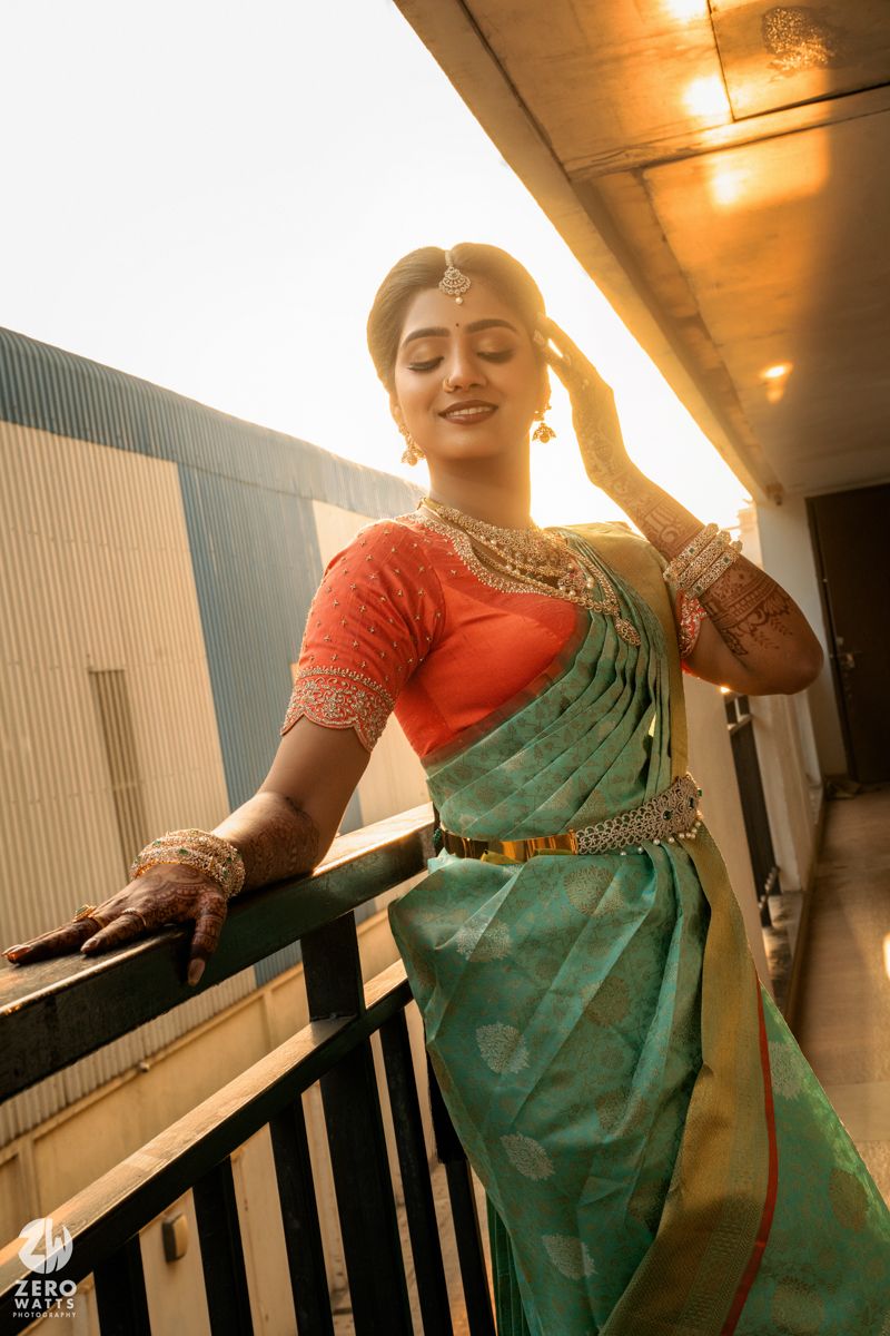 Bride in teal Banarasi saree with orange blouse and gold jewelry poses on balcony in golden sunset light.