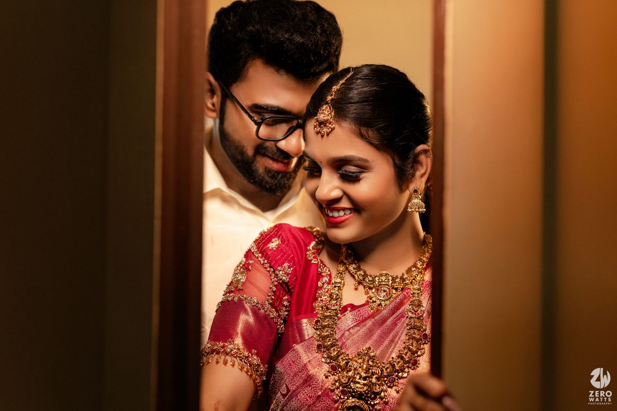 South Indian couple sharing intimate moment, bride in pink saree with gold jewelry smiling at groom.