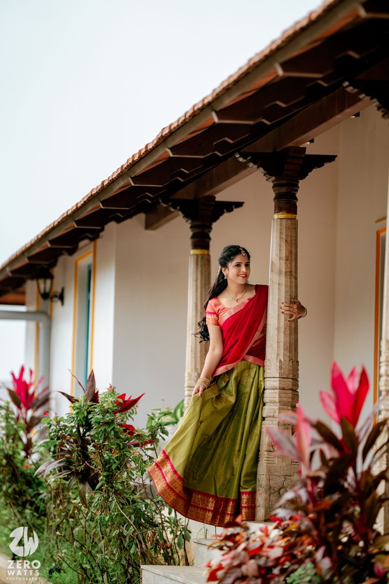 Bride in red and green silk saree posing gracefully between stone pillars of a traditional South Indian heritage home.