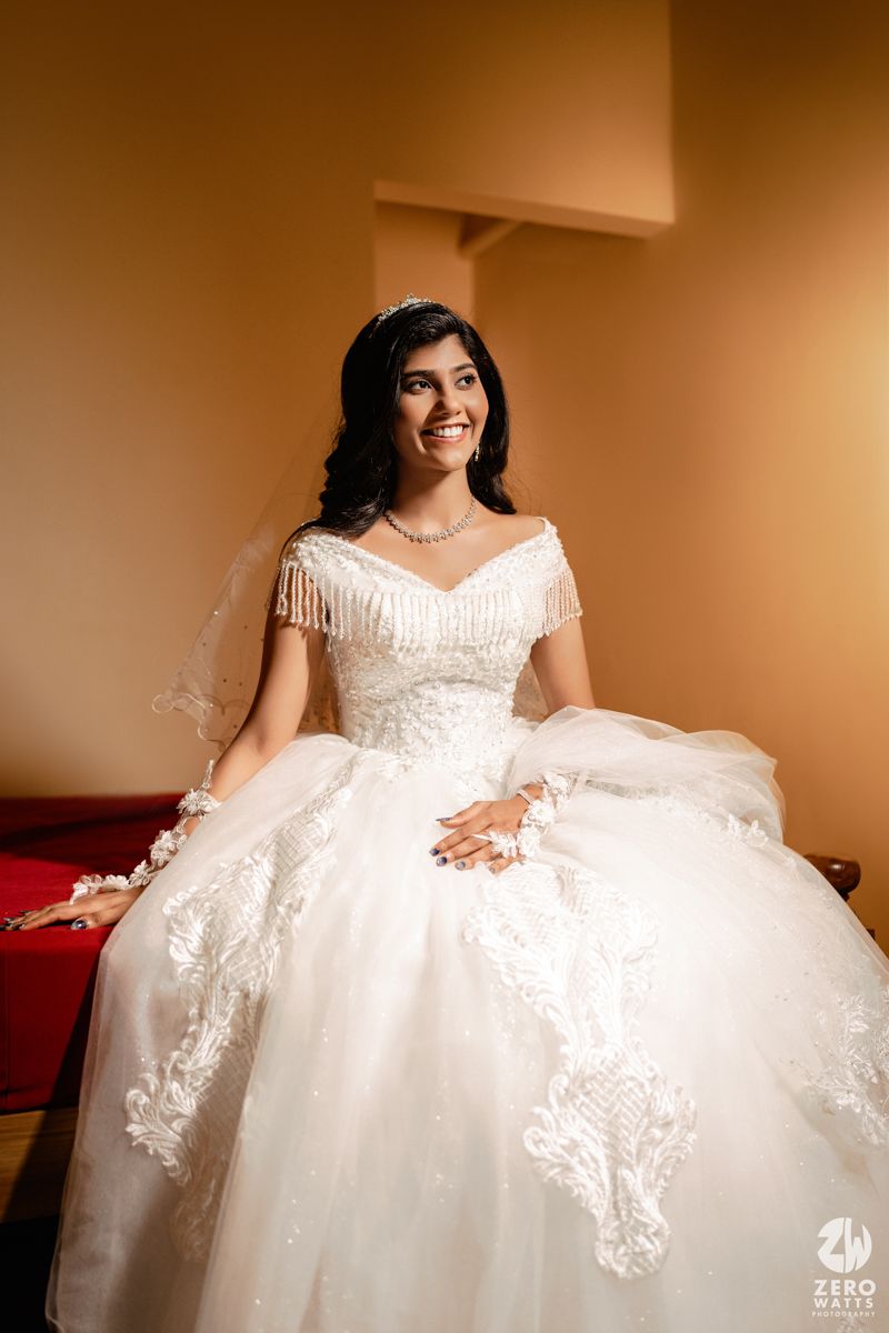 Smiling bride in white off-shoulder ball gown with fringe details and veil, seated in warm-lit room.