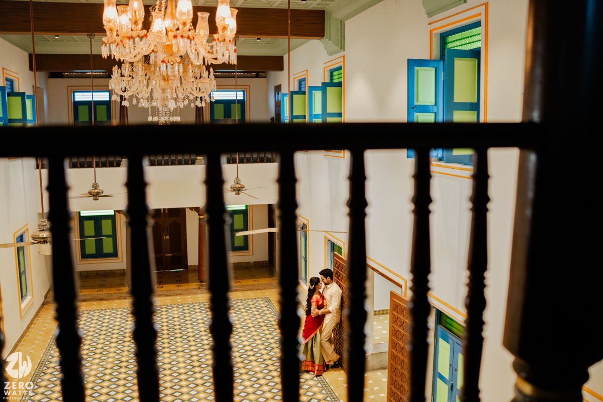 Couple in traditional Indian attire framed by ornate heritage mansion interior with chandelier and mosaic floors.