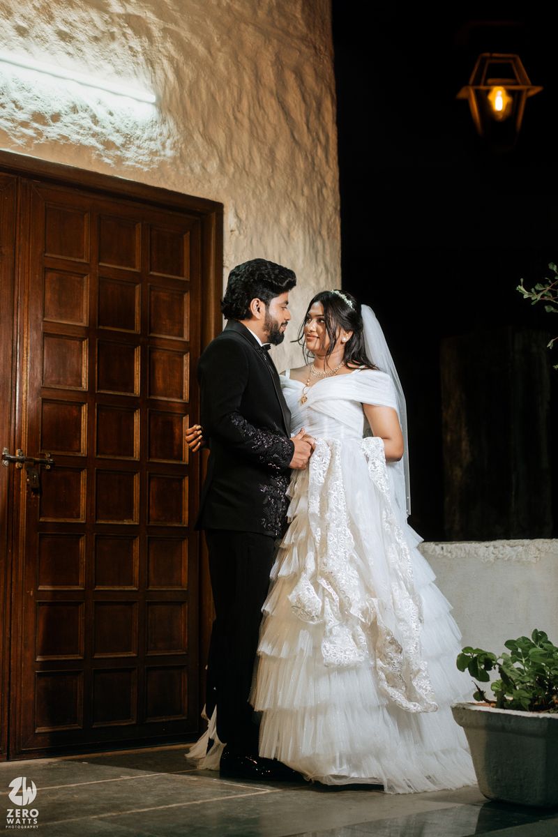 Bride in white ruffled gown and veil stands with groom in black suit near wooden door at night.