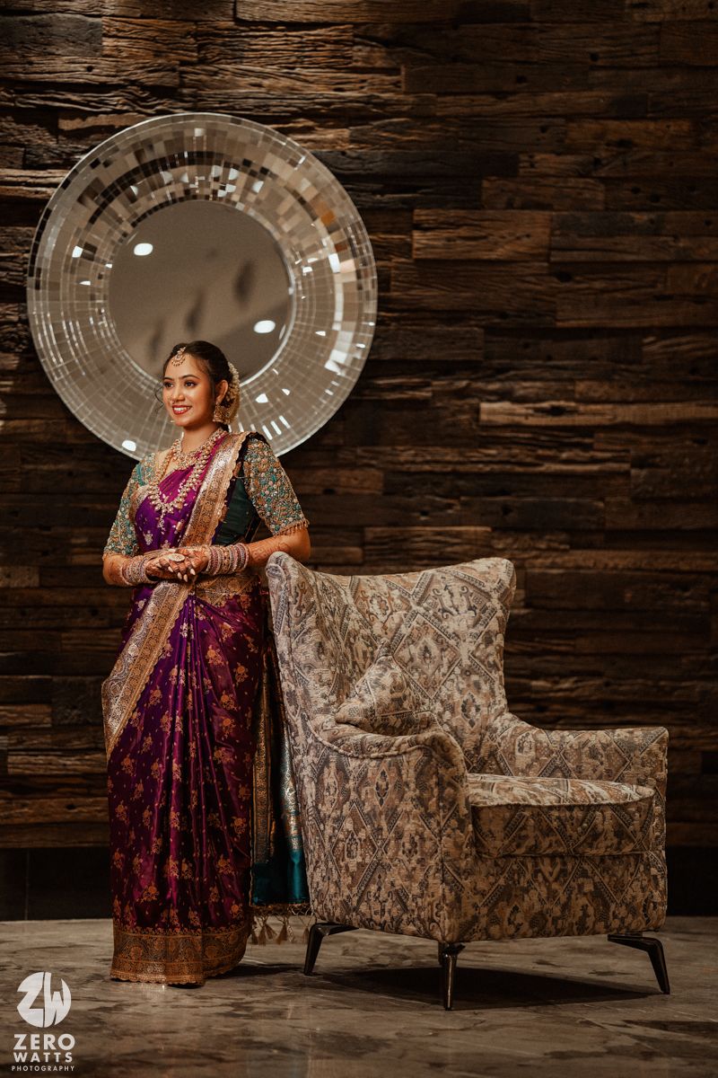 Bride in purple silk saree with green blouse poses elegantly beside ornate chair against wooden wall with round mirror.