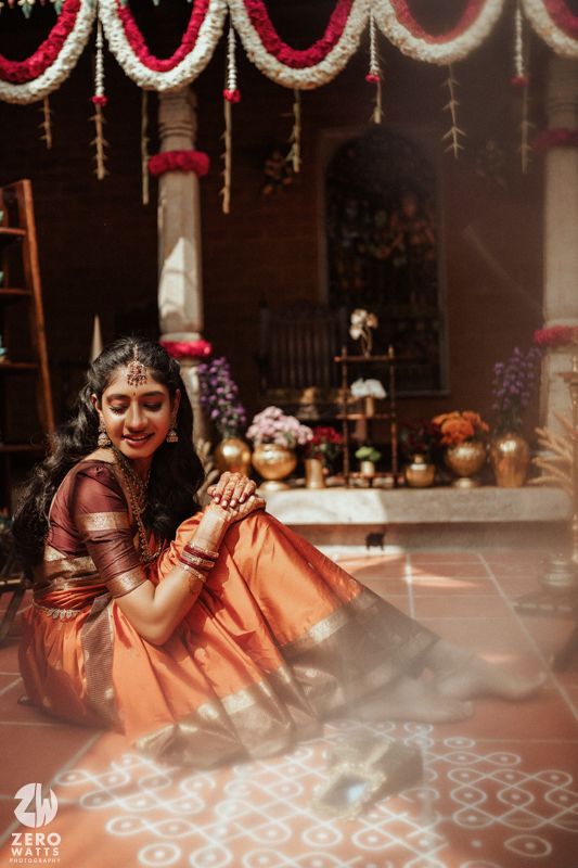 South Indian bride in orange Kanjivaram saree sitting beside kolam rangoli under floral garland decorations.