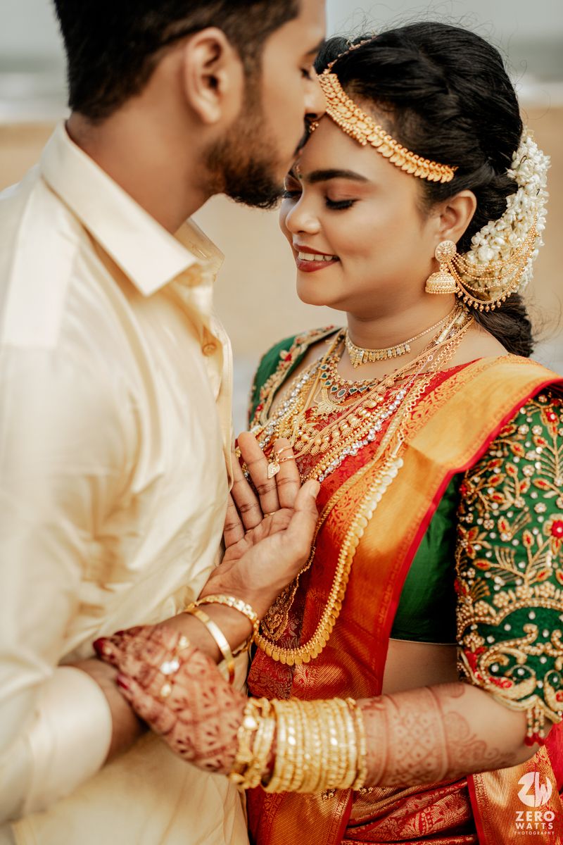 South Indian bride in red and green silk saree with gold jewelry shares romantic moment with groom in cream shirt.