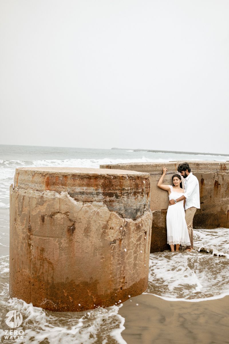 Couple embracing by rustic concrete sea barriers on a beach, woman in white dress, waves washing around their feet.