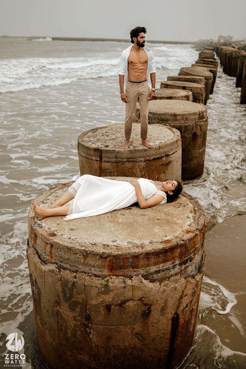 Woman in white dress lying on a concrete pier pillar while man stands behind her overlooking the ocean.