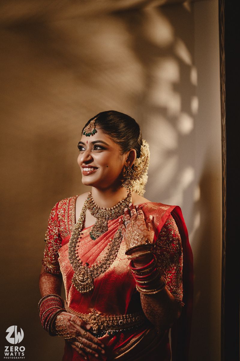 A bride in traditional South Indian wedding attire wearing a red silk saree and ornate gold jewelry poses elegantly.