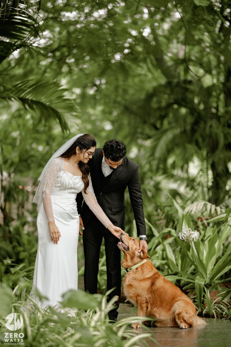 A couple in wedding attire pets their golden retriever dog in a lush tropical garden setting.